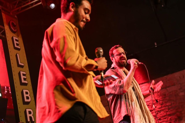 Dois cantores se apresentam juntos no palco, sob a luz vermelha.