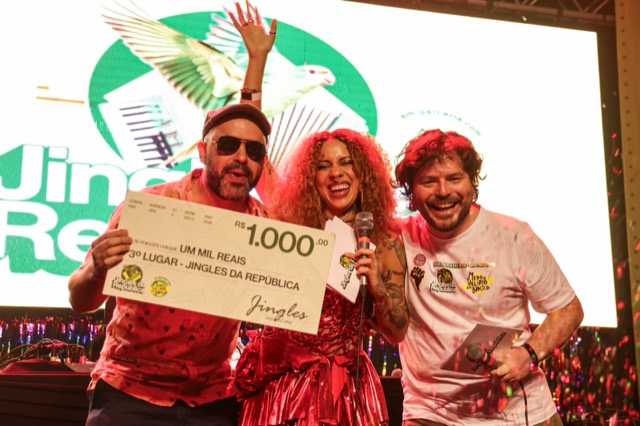 Premiado de óculos escuros ergue o cheque simbólico de R$ 1.000 entre os apresentadores.